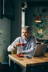 handsome gray haired man drinking coffee and working on laptop in cafeteria