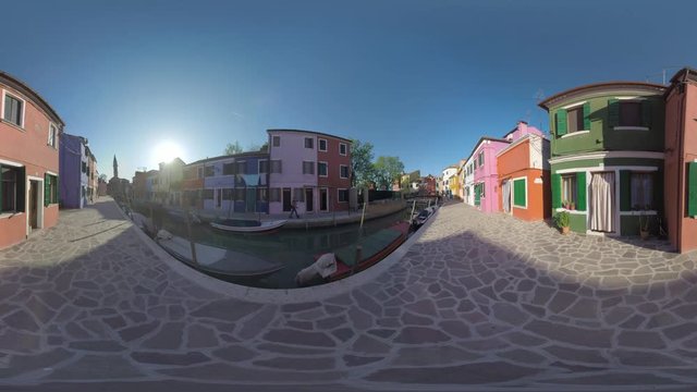 360 VR video. Burano island in Italy. Scene with paved streets and traditional colourful houses along the canal with moored boats. View with Leaning Bell Tower and bright sunshine
