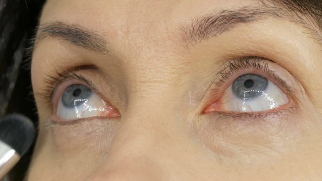 Make up artist do makeup with highlighter concealer to middle-aged woman with blue eyes. Age makeup. Close-up portrait. Apply powder with a brush