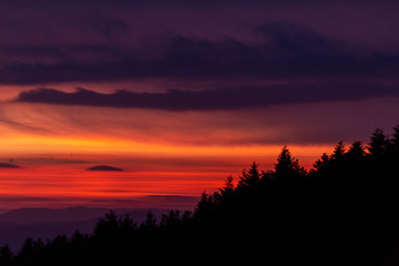 Trees silhouettes against a beautifully colored sky at dusk, with mountains layers in the background