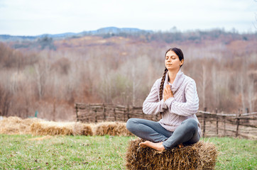 Naklejka na ściany i meble woman with long braid sitting on a hay bale meditating in a beautiful rural landscape
