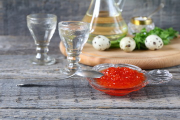 Red caviar in glassware near the glass of vodka on a gray wooden background.