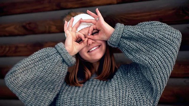 I am batman dude. Portrait of a joyful attractive girl in a sweater and a white hat. Showing I am batman signs with both raised hands and smiling broadly as if taking picture