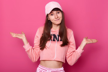 Isolated shot of attractive brunette pretty teenager, wears rose hoodie and cap, looking at camera, has long dark hair, she spreads her tanned hands, makes helpless gesture, poses on pink studio wall.