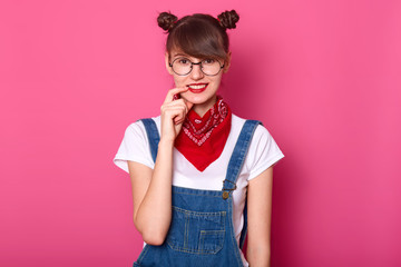 Dark haired student smiles, keeps fore finger on her lip, looks shy. Young girl wears t shirt, denim overalls with red bandana on neck. Model poses in photo studio isilated over pink background.