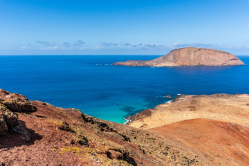 Fototapeta na wymiar Isla de Montana Clara island as seen from the top of Bermeja volcanic mountain on La Graciosa Island in Lanzarote, Spain. Colorful landscape with red rugged cliffs and turquoise sea waters.