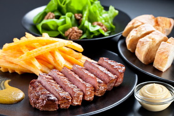 Appetising steak slices served with fries , green salad and baguette on a black plate.