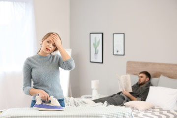 Tired housewife ironing clothes while her lazy husband lying on bed