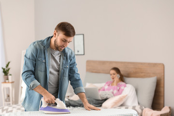 Househusband ironing clothes while his wife working in bed