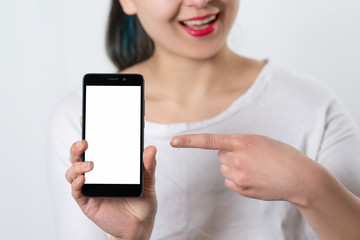 a young smiling woman holds a smartphone vertically with a white screen with a place for copyspace and points at him the finger of the other hand. on white background