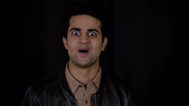 Young Indian man is completely shocked and surprised at some great news from off camera as his expression changes and smiles and eyes widen and he fists the air with both hands 