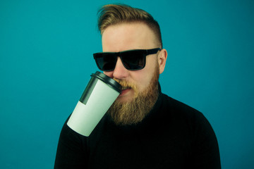 young fashionable handsome guy in a black turtleneck, holding a white plastic Cup of coffee on a blue background, a man drinking coffee