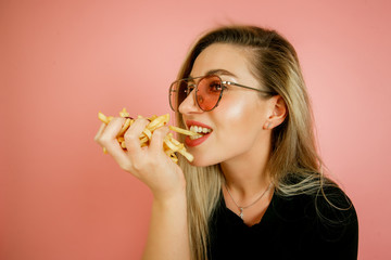 young beautiful blonde girl model with appetite eating fast food, French fries holding in hand, on a pink background