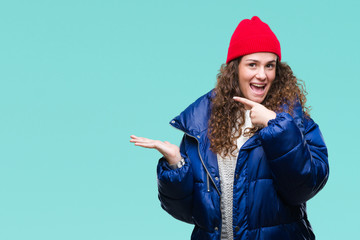 Beautiful young brunette curly hair girl wearing winter coat, wool cap and sweater over isolated background amazed and smiling to the camera while presenting with hand and pointing with finger.