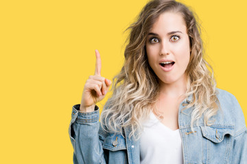 Beautiful young blonde woman wearing denim jacket over isolated background pointing finger up with successful idea. Exited and happy. Number one.