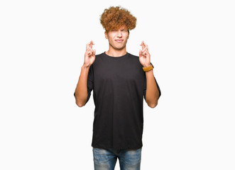 Young handsome man with afro hair wearing black t-shirt smiling crossing fingers with hope and eyes closed. Luck and superstitious concept.