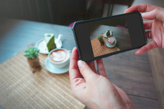 Hands taking photo or snap and share on social media by smartphone  to take a photo of coffee break latte mocha coffee with art foam matcha cake and cactus pot on a map and wooden table background 