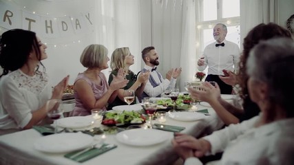 A big family sitting at a table on a indoor birthday party, a senior man giving a speech.