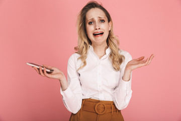 Portrait of resentful blond woman 30s in stylish outfit crying while holding and using smartphone