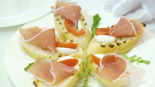 A cook decorates a ceramic plate of sandwiches made of cured meat, cherry tomatoes and quail eggs, laying green cilantro leaves between them. Serving of dishes. Close-up.