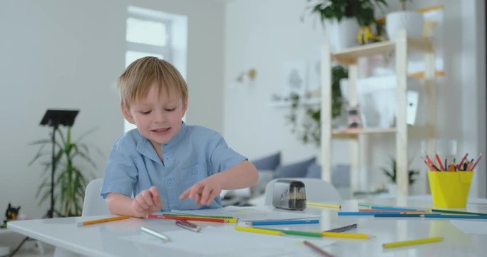 Smiling boy in blue shirt draws on paper with a pencil while sitting at the table in the living room