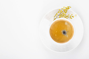 White cup of coffee on a white table with yellow mimosa flowers. Minimalism. Top view, flat lay