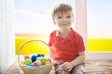 Positive kid holding basket full of easter eggs after egg hunt enjoying and sitting on the windowsill against background of a spring green field at sunset or dawn