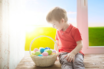 Cheerful smiling boy holding basket full of colorful easter eggs and sitting on the windowsill against the background of spring green field at sunset or dawn