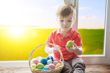 Positive kid holding basket full of easter eggs after egg hunt enjoying and sitting on the windowsill against background of a spring green field at sunset or dawn