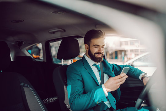 Young Caucasian successful bearded businessman in turquoise suit and with toothy smile sitting in the car and using smart phone. The expert in anything was once a beginner.