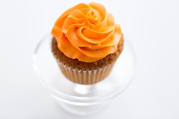 food, pastry and sweets concept - cupcake with orange buttercream frosting on glass confectionery stand over white background