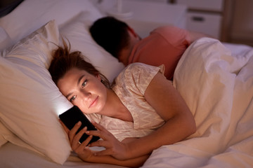 technology, internet addiction and cheat concept - woman using smartphone at night while her boyfriend is sleeping - obrazy, fototapety, plakaty
