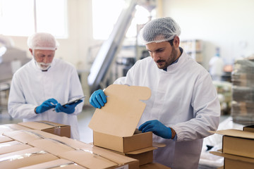 Young Caucasian employee in sterile uniform packing goods in boxes. In background supervisor...