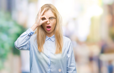 Young beautiful blonde business woman wearing glasses over isolated background doing ok gesture shocked with surprised face, eye looking through fingers. Unbelieving expression.