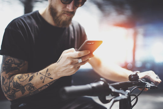 Bearded tattooed man in sunglasses using mobile phone for send text message after riding by electric scooter in the city