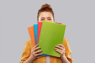 education, school and knowledge concept - red haired teenage student girl hiding behind notebooks over grey background