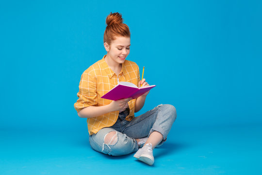 education, high school, inspiration and people concept - smiling red haired teenage girl in checkered shirt and torn jeans with diary or notebook and pencil over grey background