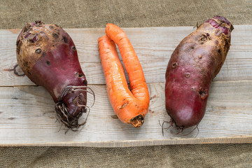 Three non-standard ugly vegetables are lying on grey wooden planks on burlap. 