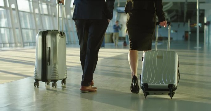 Back view on the Caucasian businessman and businesswoman walking the airport hall and carrying their suitcases on the wheels before the departure to the working trip. Rear. Camera moving from feet up.