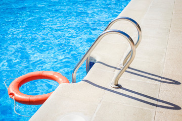 Closeup view of the swimming pool edge with staircase and a red ring life buoy floating on the blue water during a sunny summer day