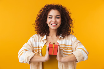 Beautiful young curly girl posing isolated over yellow wall background holding present gift box.