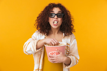 Cute beautiful young curly girl posing isolated over yellow wall background wearing glasses eat popcorn watch film.