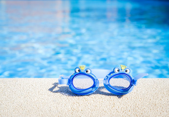 Closeup view of a boy blue swimming goggles on the pool edge with a big blue water copy space during a sunny summer day