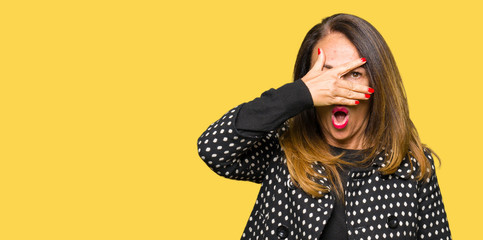 Beautiful middle age woman wearing fashion jacket peeking in shock covering face and eyes with hand, looking through fingers with embarrassed expression.