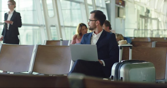 Portrait shot of the Caucasian attractive man in glasses and business style sitting in the airport and typing on the laptop computer while waiting for his flight.