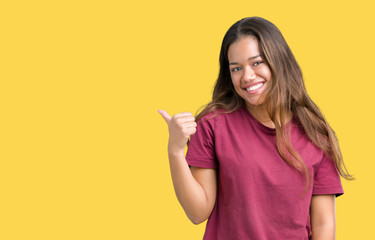 Young beautiful brunette woman over isolated background smiling with happy face looking and pointing to the side with thumb up.