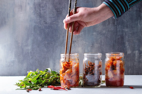 Korean traditional fermented appetizer kimchi cabbage and radish salad, fish snack served in glass jars with Vietnamese oregano and chili peppers over grey blue table. Chopsticks in mans hands.