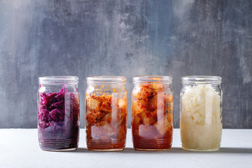 Variety of fermented food korean traditional kimchi cabbage and radish salad, white and red sauerkraut in glass jars in row over grey blue table. - obrazy, fototapety, plakaty