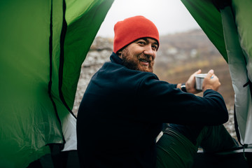Portrait of a view from tent entrance of young traveler man smiling and relaxing in mountains near...