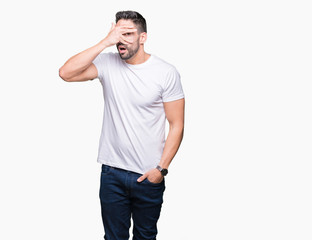 Young man wearing casual white t-shirt over isolated background peeking in shock covering face and eyes with hand, looking through fingers with embarrassed expression.